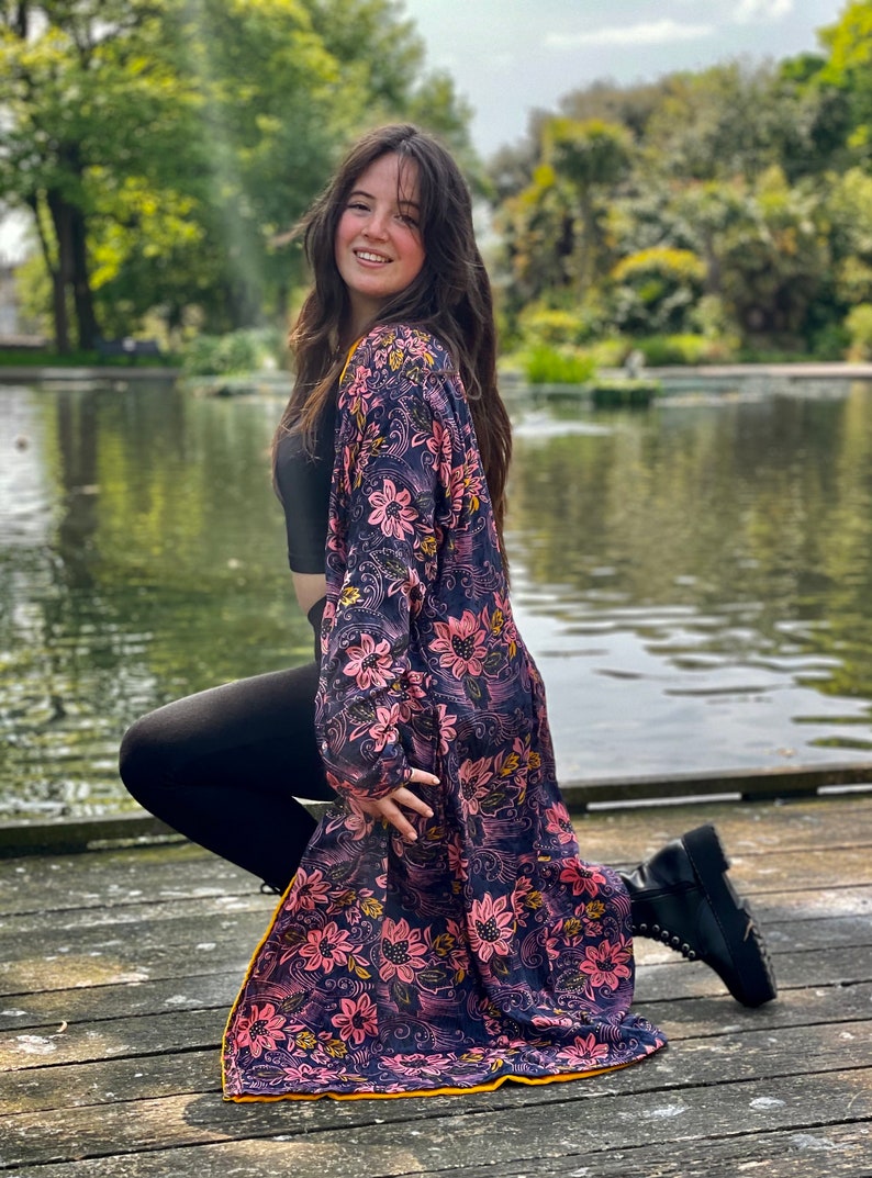 K&ouml;nnte beinhalten: Eine Frau tr&auml;gt einen langen, floral gemusterten Kimono mit einem schwarzen Crop-Top und schwarzen Leggings. Sie kniet auf einem Holzsteg mit einem Teich im Hintergrund.