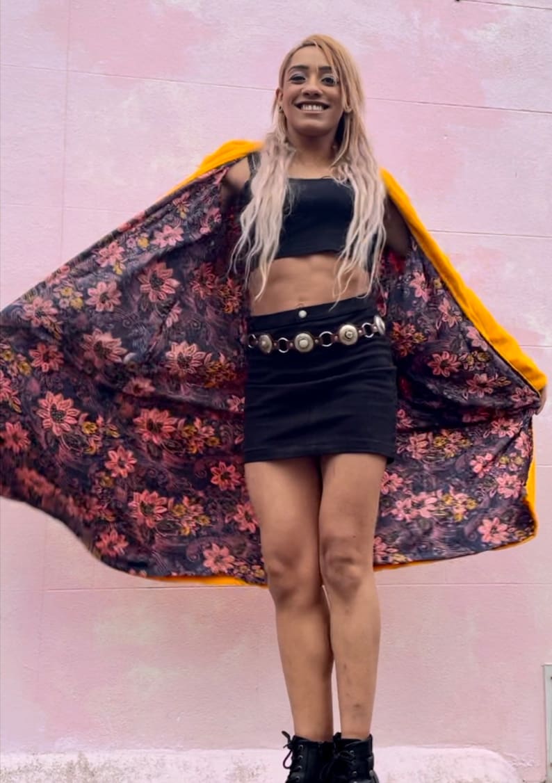 K&ouml;nnte beinhalten: Eine Person tr&auml;gt einen gelben Kunstpelzmantel mit einem floralen Futter. Der Mantel ist offen und die Person tr&auml;gt ein schwarzes Crop-Top und einen schwarzen Minirock mit einem silbernen G&uuml;rtel. Die Person steht vor einer pinkfarbenen Wand.