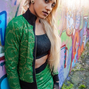 May include: A woman wearing a green and gold patterned bomber jacket with a black zipper and a matching skirt. She is standing in front of a graffiti wall.