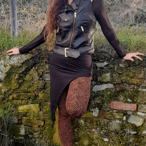 May include: A woman wearing a black leather jacket, a black skirt, and brown patterned leggings. She is standing in front of a stone wall.