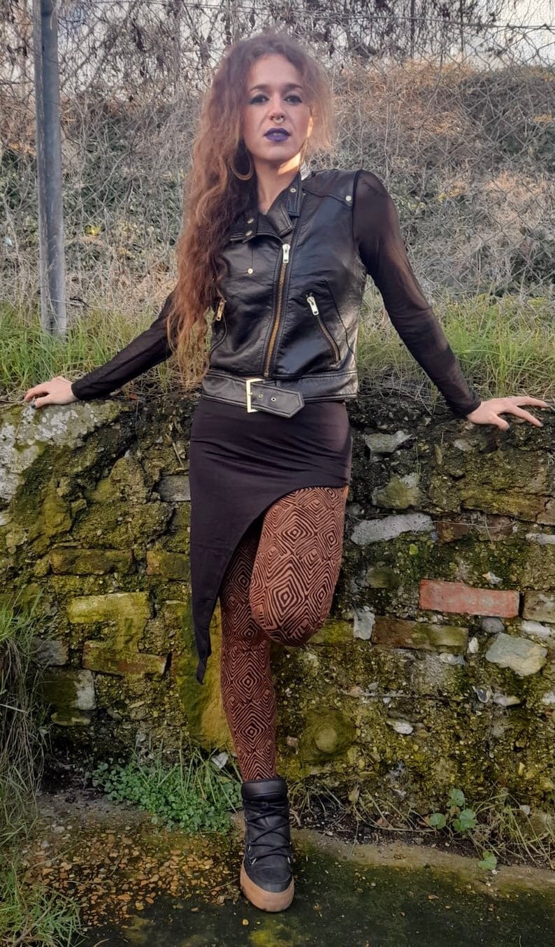 May include: A woman wearing a black leather jacket, a black skirt, and brown patterned leggings. She is standing in front of a stone wall.