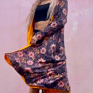 K&ouml;nnte beinhalten: Ein langer Cardigan mit floralem Muster und gelbem Futter. Der Cardigan wird &uuml;ber einem schwarzen Crop-Top und einer schwarzen Hose getragen. Die Person, die den Cardigan tr&auml;gt, l&auml;chelt und schaut in die Kamera.