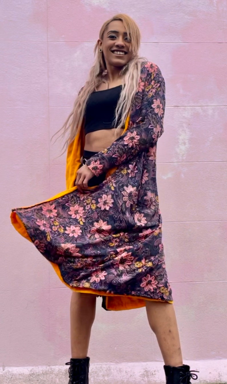 K&ouml;nnte beinhalten: Ein langer Cardigan mit floralem Muster und gelbem Futter. Der Cardigan wird &uuml;ber einem schwarzen Crop-Top und einer schwarzen Hose getragen. Die Person, die den Cardigan tr&auml;gt, l&auml;chelt und schaut in die Kamera.
