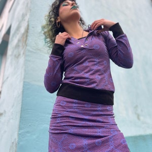 Puede incluir: Una blusa de manga larga de color morado y negro con un patrón geométrico y una banda negra. La blusa tiene capucha y cierre de botón. La modelo lleva una falda morada a juego con un patrón geométrico.