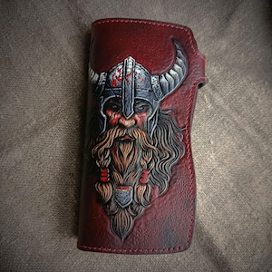 Puede incluir: Cartera de cuero burdeos con imagen en relieve de un guerrero vikingo. El guerrero tiene una larga barba, un casco con cuernos y una expresión severa. La cartera es rectangular y parece estar hecha a mano. Mide aproximadamente 10 cm x 20 cm.