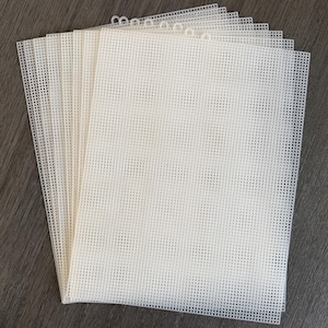 May include: A stack of off-white plastic canvas sheets. Each sheet has a grid of evenly spaced square holes, and a series of small loops along one edge. The sheets are stacked on a wooden surface.
