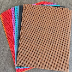 May include: A stack of colorful plastic canvas sheets in various colors, including orange, red, pink, teal, gray, and brown. Each sheet has a grid pattern of square holes, ideal for needlepoint and cross-stitch projects.