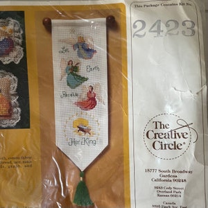 May include: A cross-stitch kit featuring a religious banner with angels and the text "Let Earth Receive Her King." The banner is white with a green tassel and wooden dowels. The kit includes the number 2423 and the logo for The Creative Circle.