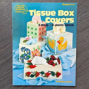 May include: A vintage booklet titled "Tissue Box Covers" featuring eight designs in plastic canvas. The cover displays several finished tissue box covers with floral and duck designs. The booklet is by Barbara Hunter and published by the American School of Needlework.