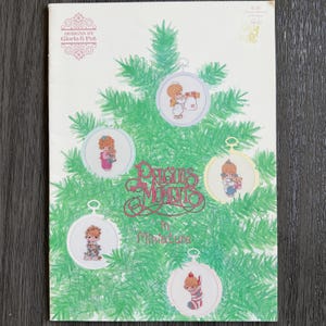 May include: A vintage craft pattern book titled "Precious Moments in Miniature" featuring cross-stitch designs. The cover shows a Christmas tree with ornaments, each depicting a Precious Moments character. The book's cover is cream-colored with green and pink accents.