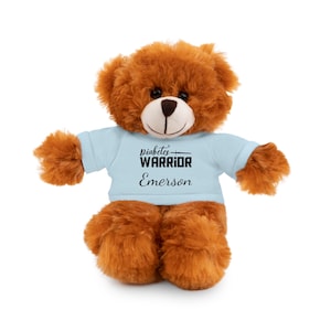 May include: A brown teddy bear wearing a light blue t-shirt. The shirt features the words "Diabetes WARRIOR" and the name "Emerson" printed in black. The bear has a light brown snout and dark eyes.