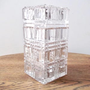 Può includere: Un vaso di vetro trasparente con un motivo geometrico. Il vaso è quadrato e ha una superficie testurizzata.