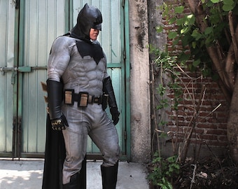 May include: A full-body Batman costume featuring a grey textured bodysuit with muscle definition, a black cowl, a black cape, black gauntlets, black boots with metallic toe caps, and a utility belt with rectangular pouches in black and gold tones.