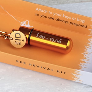 May include: A gold metal keychain with a small, round container attached to it. The container is engraved with the text "Leo - 19.06". The keychain also has a round tag with the text "BEE" and a bee symbol. The keychain is packaged in a box with the text "BEE REVIVAL KIT".