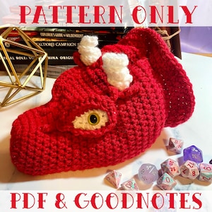May include: Red crocheted dragon dice bag with white horns and a yellow eye. The bag is sitting on a white surface with several multi-colored dice scattered around it. The text "PDF & GOODNOTES" is printed at the bottom of the image.
