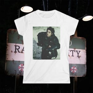 May include: A white short-sleeved t-shirt featuring a central rectangular graphic print of a person in a dark jacket holding a black medical bag with a white cross symbol. The background of the print shows a muted, light-coloured outdoor setting.