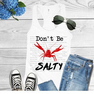 K&ouml;nnte beinhalten: Ein wei&szlig;es Tanktop mit einer roten Krebsgrafik und dem Text "Don't Be Salty". Das Tanktop liegt auf einer Holzoberfl&auml;che mit einer blauen Jeans, einem Paar schwarzen Turnschuhen und einer Sonnenbrille.