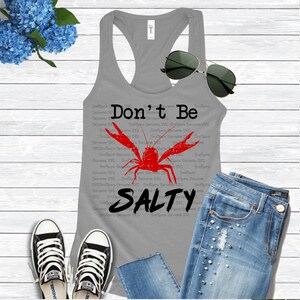K&ouml;nnte beinhalten: Ein graues Tanktop mit dem Text "Don't Be Salty" und einer roten Krebsgrafik. Das Tanktop ist mit einer blauen Jeans und wei&szlig;en Turnschuhen gestylt.