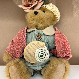 Pode incluir: Um urso de peluche castanho que veste um vestido de veludo cotelê azul, um casaco de malha rosa, um chapéu de crochê branco e uma rosa de tecido rosa. O urso está sentado com as patas dobradas à sua frente. O urso tem uma etiqueta que diz "Boyds Bears Bearwear Presents Bailey the Bear".