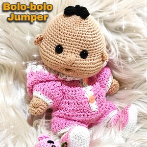 Puede incluir: Una muñeca de crochet vestida con un mono rosa y blanco con un botón. La muñeca tiene el pelo negro y los ojos marrones. La muñeca está sentada sobre una superficie blanca y esponjosa con un juguete de crochet rosa.