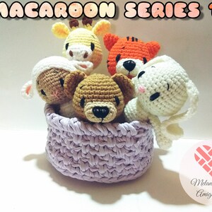 May include: A set of five crocheted animal plushies in a lavender basket. The plushies include a giraffe, tiger, bear, bunny, and lamb. The plushies are all in a sitting position and have embroidered faces. The basket is made of a crocheted material and has a light lavender colour. The image includes the text "Macaroon Series 1" and "Melani Koa Amigurumi".