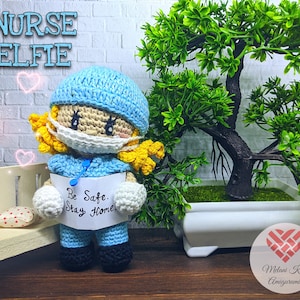 May include: A crocheted elf doll dressed as a nurse, wearing a blue scrub top, a white face mask, and a blue surgical cap. The doll is holding a sign that says "Be Safe. Stay Home." The doll is sitting on a wooden bench in front of a potted plant.