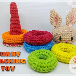 Può includere: Un giocattolo impilabile di coniglio all'uncinetto con anelli colorati. Gli anelli sono rosso, arancione, blu, giallo e verde. Il coniglio è beige con occhi neri e un naso rosa. Il giocattolo è etichettato "Bunny Stacking Toy".