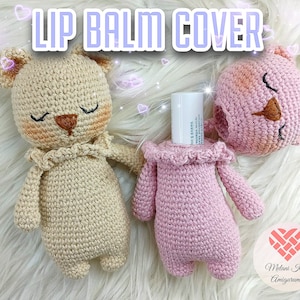 May include: Two crocheted stuffed toys, one beige and one pink, designed to hold lip balm tubes. The beige toy has a brown nose and closed eyes. The pink toy has a brown nose and closed eyes. The pink toy is holding a white lip balm tube. The text "Lip Balm Cover" is displayed at the top of the image.