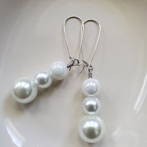 May include: A pair of drop earrings with white, pearlescent beads. The beads are arranged in a descending size order. The earrings have long, silver-coloured ear wires.