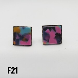 May include: A pair of square earrings with a pink, blue, black, and yellow marbled design. The earrings have a bronze-colored metal frame.