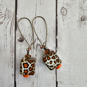 May include: A pair of dangle earrings with a leopard print design. The earrings are made of metal and have a brown and orange colour scheme.