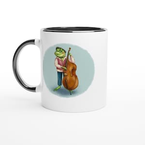 May include: A white ceramic mug with a black interior and handle, featuring a graphic illustration of a green frog wearing a vest and cap while playing a large brown upright bass, set against a soft blue circular background on the mug surface.