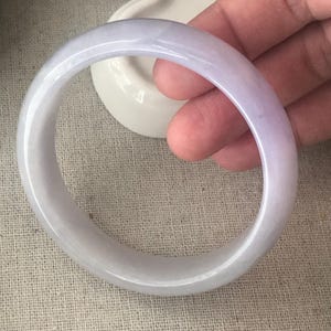 May include: A pale purple jade bangle bracelet. The smooth, polished surface of the bracelet reflects light, showcasing a subtle gradient of colour. The bracelet is held against a neutral background, highlighting its elegant design.