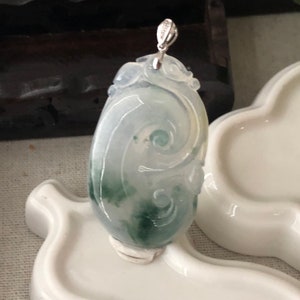 May include: An oval jade pendant with a swirling pattern in green and white. It has a silver bail. The pendant is displayed on a white ceramic surface. The jade's design is carved.