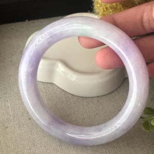 May include: A polished jade bangle bracelet with a circular design. The bracelet displays a colour gradient, shifting from a pale lavender to a deeper purple. The jade is smooth and translucent, with a glossy finish. The bangle is held against a white background.