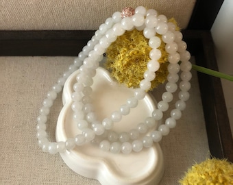 May include: A white beaded necklace with a rose gold clasp. The necklace is draped over a white, decorative dish. The beads are round and appear to be made of a smooth, polished material. The background includes a beige fabric and yellow floral accents.
