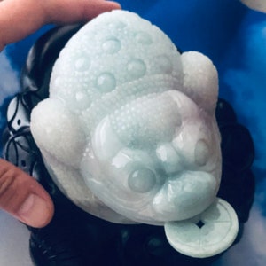 May include: A light blue and white jade frog sculpture with a coin in its mouth. The frog has a textured surface and is resting on a black base. The sculpture is held by a hand.