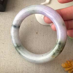 May include: A light lavender jade bangle with green and white swirls. The bangle is smooth and polished.