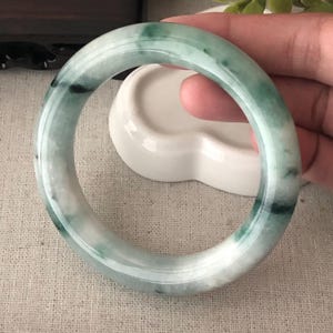 May include: A jade bangle bracelet with a smooth, polished surface. The bracelet features a blend of white and green hues, with darker green inclusions. The circular design is a classic jewelry style.