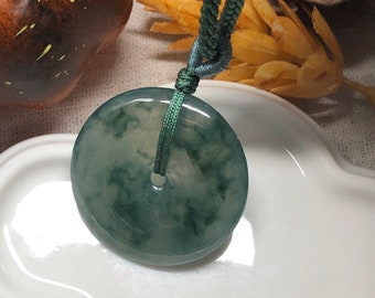 May include: A jade pendant with a circular design and a green cord. The pendant has a translucent appearance with green and white marbling. The cord is a dark green color and is knotted at the top of the pendant.