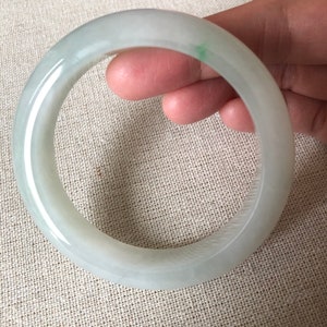 May include: A pale green jade bangle. The smooth, circular bracelet has a translucent appearance with subtle colour variations. A small area of darker green is visible. The bracelet is held by a hand.