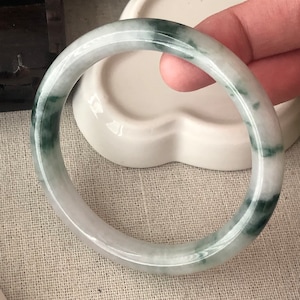 May include: A jade bangle bracelet with a white and green colour scheme. The bangle is smooth and polished, with a subtle swirl pattern.