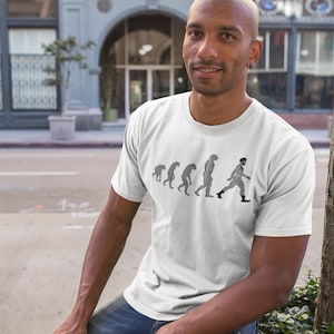 May include: A white t-shirt featuring a black and white graphic depicting the evolution of man, culminating in a man wearing leather trousers and boots. The man is smiling and wearing a white t-shirt.