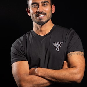 Peut inclure: T-shirt noir à col en V avec un petit motif brodé sur la poitrine. Le motif est de forme triangulaire avec un motif floral. La personne sur l'image a les bras croisés et sourit. Le fond est noir.