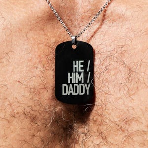 May include: A silver chain necklace with a black dog tag pendant. The dog tag has the words "HE/HIM/DADDY" printed in white.