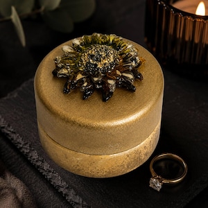 May include: A round, metallic gold jewelry box with a textured finish, featuring a decorative, dark-toned, multi-petaled flower ornament on the lid. A simple gold band ring with a single clear stone sits beside the container.