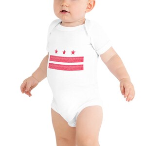 May include: White baby bodysuit with a red design of three stars above two horizontal stripes. The graphic is centered on the front of the onesie. The baby bodysuit is made of soft material.