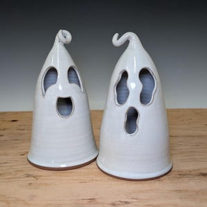 May include: Two white ceramic ghost figurines with glossy finishes. Each ghost features a pointed top with a small curl, two hollow eye openings, and one hollow mouth opening. The figures have a smooth, rounded base and a visible brown clay rim.