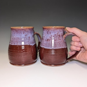 May include: Two handmade ceramic mugs. The mugs have a two-tone glaze, with a deep burgundy base and a speckled purple and blue top. Each mug has a large handle and a ribbed texture on the lower half.