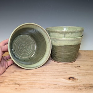May include: Handmade ceramic bowls in shades of green. The bowls have a spiral design on the inside and a textured exterior. The bowls are stacked on a wooden surface. The bowls are suitable for serving food or as decorative items.
